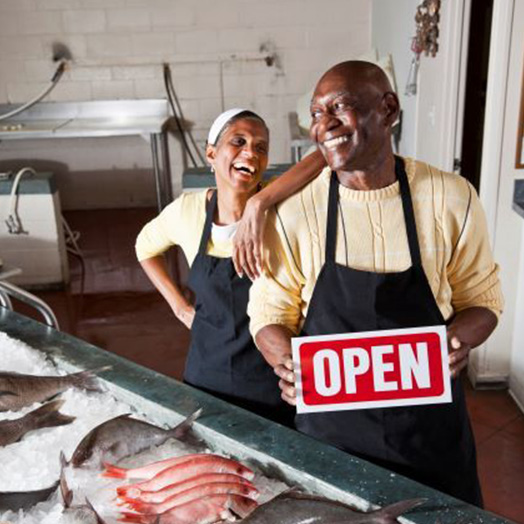 A man and a woman in a fish market smiling and holding an open for business sign.