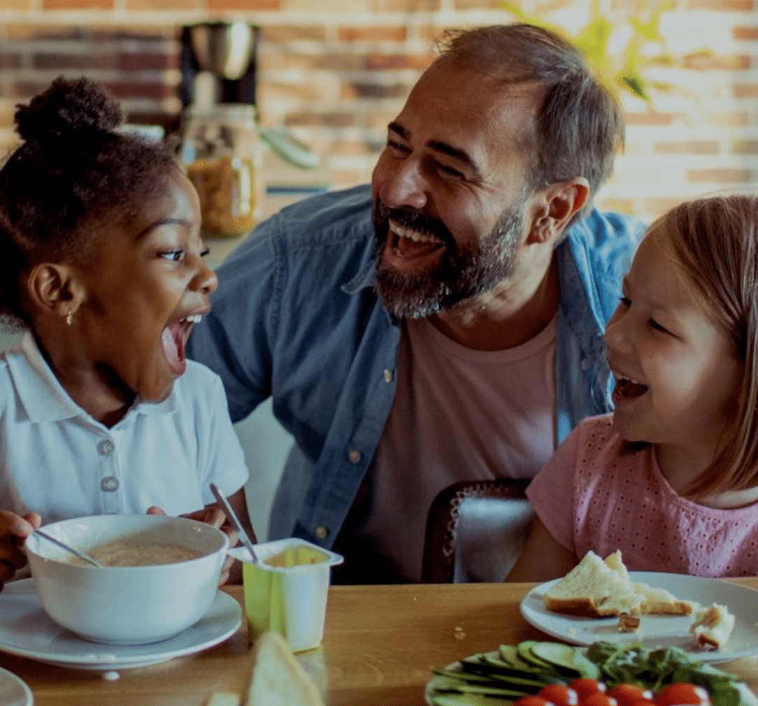 Hombre riéndose con su hija y su hijastra en la mesa del desayuno.