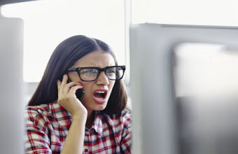Mujer joven con expresión enojada hablando por un teléfono celular mientras mira un monitor de computadora