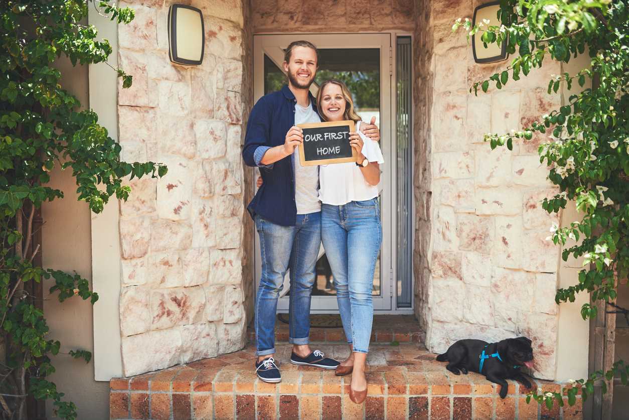New homeowners standing on porch with sign reading Our First Home. A small black dog lays near their feet.