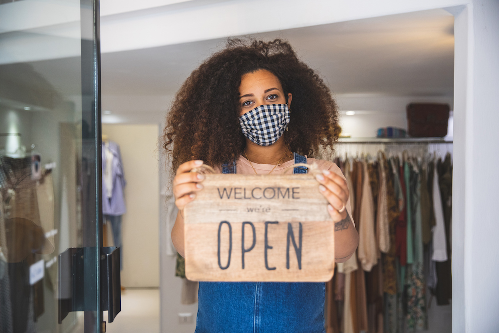 Woman wearing a mask holding Open sign in a small business boutique