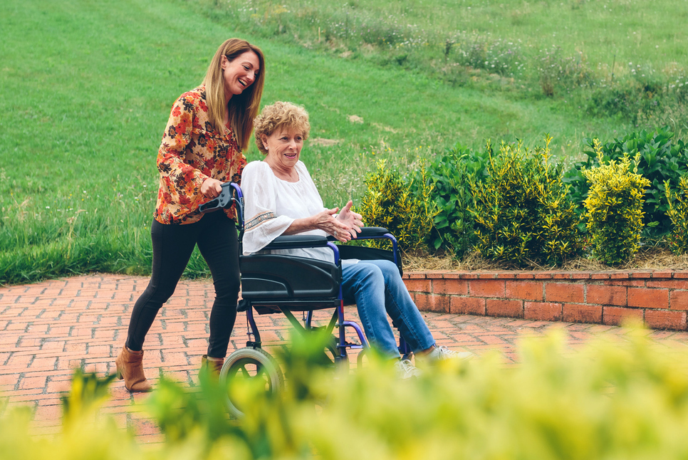 Mujer joven empujando a su madre en silla de ruedas por el jardín