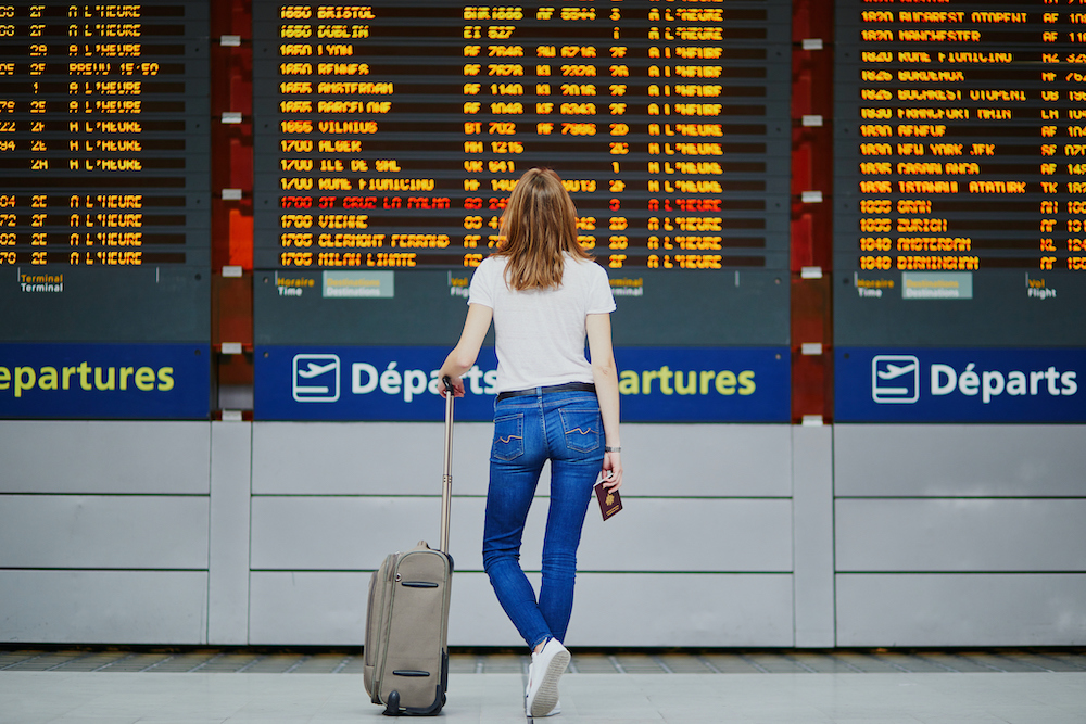 Young woman in international airport with luggage and passport looking at flight info display