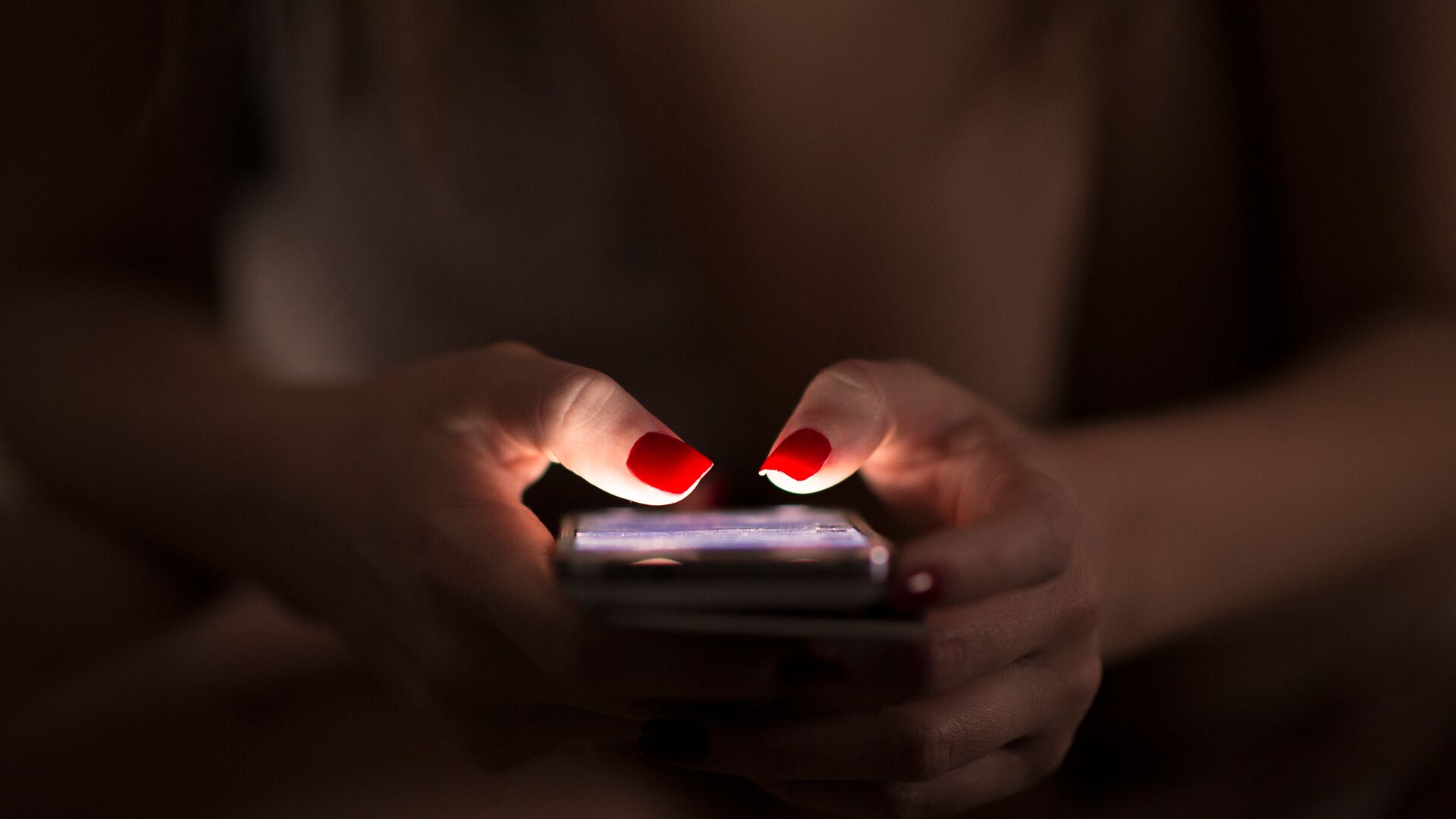 Una mujer que usa un teléfono celular en la oscuridad. Sus uñas están pintadas de rojo brillante.