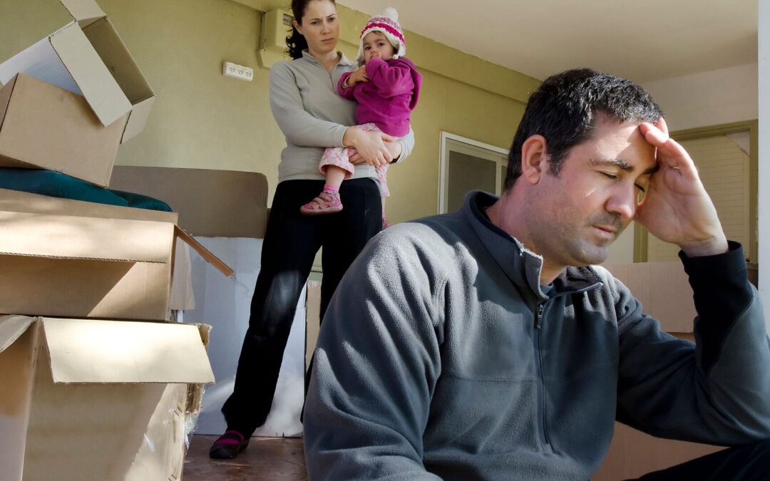 Stressed man sitting down as a woman holding a young child look on in a house being packed for a move.