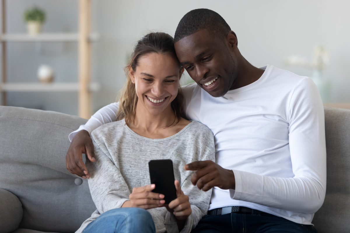 Feliz pareja sentada en un sofá y mirando un acuerdo prenupcial en un teléfono inteligente