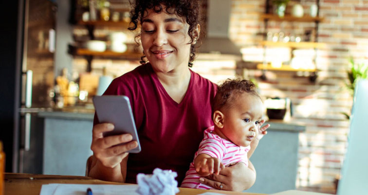 Madre con su bebé en brazos mientras ve el smartphone en su cocina