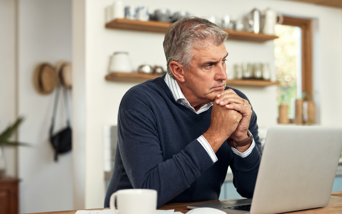 Un hombre piensa en los impuestos de su patrimonio mientras está sentado en la mesa de su casa junto a un portátil y una taza de café