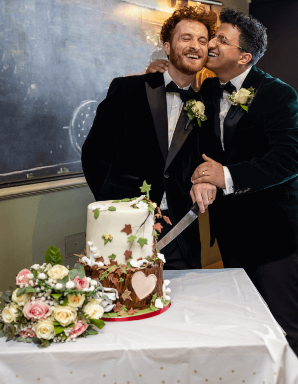 2 newly married gay men cutting their wedding cake.. One man is kissing the cheek of the other groom.