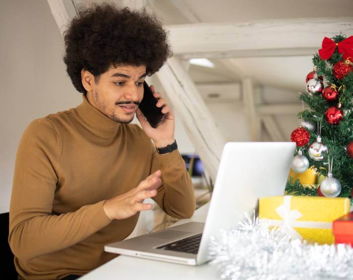 Hombre estresado hablando por teléfono y mirando la pantalla de su portátil con la esperanza de no haber sido estafado. En el fondo se ve un árbol de Navidad.