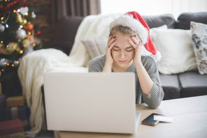 Mujer disgustada con un gorro de Papá Noel sentada en el suelo delante de un ordenador portátil sobre una mesa de centro con un árbol de Navidad de fondo.