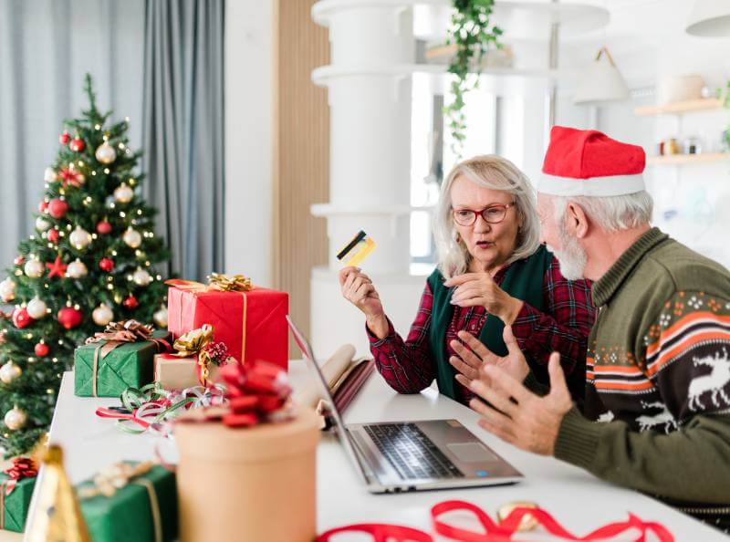 Un hombre y una mujer mayores durante la temporada navideña sentados frente a una computadora portátil. La mujer tiene una tarjeta de crédito para una compra en línea. Cerca hay un árbol de Navidad decorado.