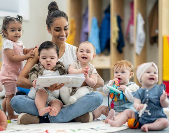 Childcare worker surrounded by babies while sitting on daycare facility floor.
