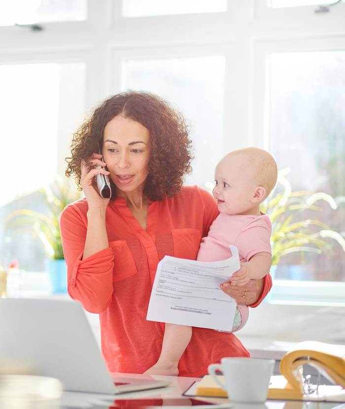 Madre con un bebé en brazos mientras mira un contrato de guardería y habla por teléfono.